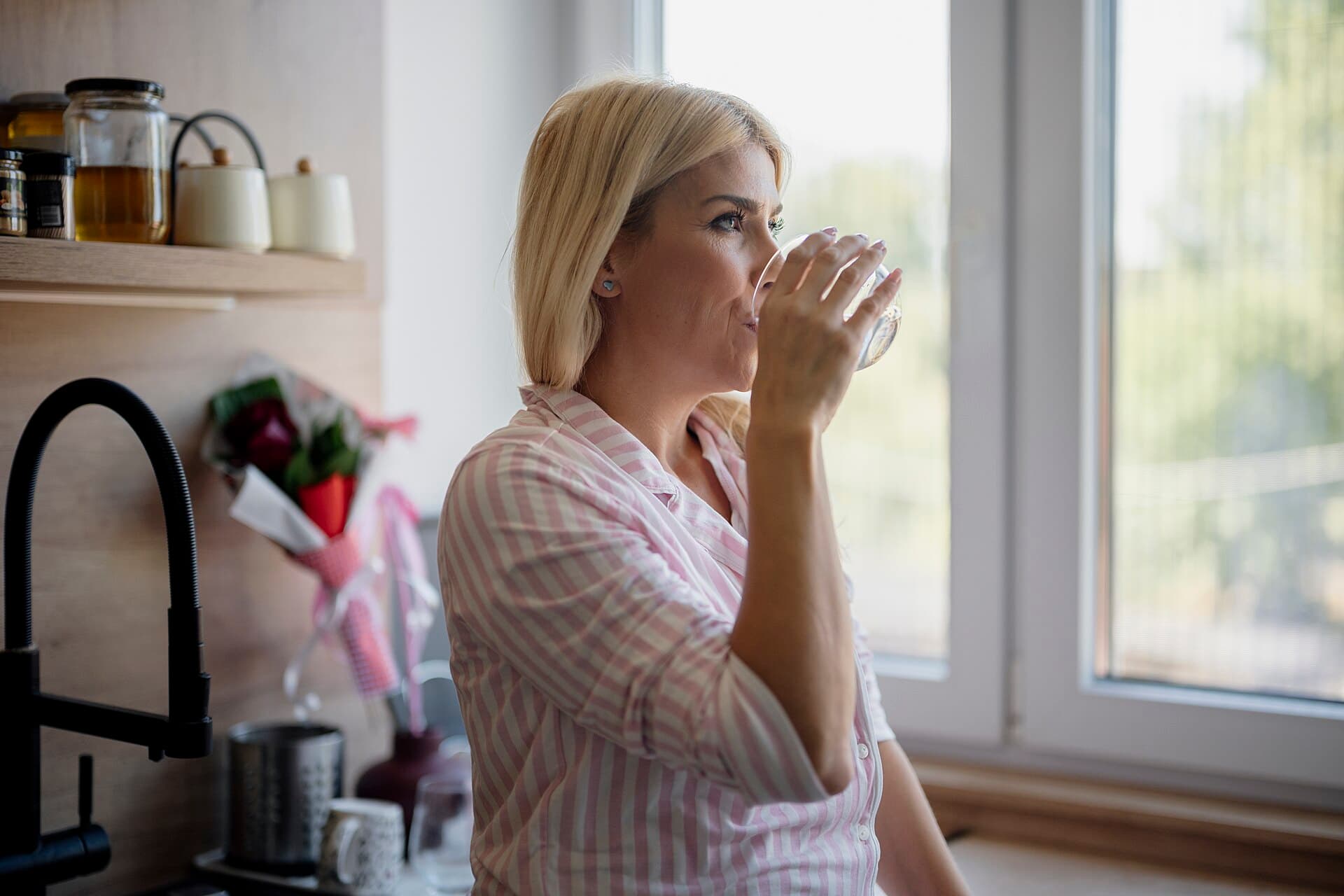 A woman drinks a glass of water in a kitchen in the morning
